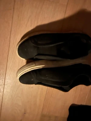 Svarta slip-on sneakers - Säljer ett par svarta slip-on sneakers med vit sula. Skorna har en enkel och clean design utan snörning, vilket gör dem lätta att ta på och av. Ovandelen verkar vara i textil eller mockaimitation och sulan är i gummi. Perfekta för en avslappnad streetstil.