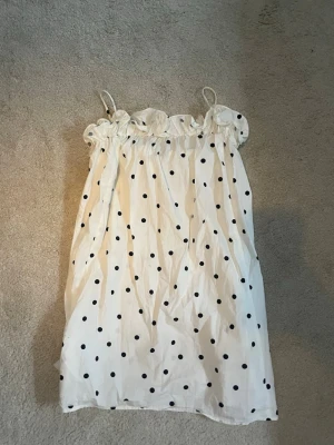 Vit prickig kort klänning - Söt vit kort klänning med svarta prickar och tunna axelband. Klänningen har volangdetalj upptill och är perfekt för varma dagar. Tillverkad i ett lätt och luftigt material som känns skönt mot huden.