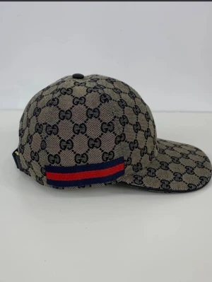 Gucci keps med GG-mönster - Snygg keps från Gucci i beige med klassiskt GG-monogram över hela kepsen. Har en böjd skärm och en röd-blå rand på sidan. Tillverkad i canvasmaterial med justerbar rem bak. Perfekt för dig som vill sticka ut med en lyxig accessoar.vill bli av med snabbt.