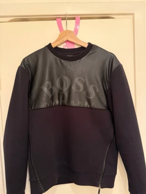 Svart sweatshirt från Hugo Boss - Snygg svart sweatshirt från Hugo Boss med läderliknande panel över bröstet och stort BOSS-tryck. Tröjan har rund halsringning, långa ärmar och dragkedjedetalj nedtill. Perfekt för dig som gillar streetwear och exklusiv känsla.