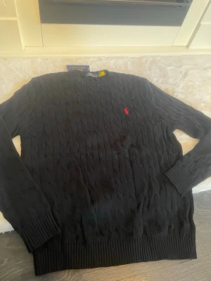 Svart kabelstickad tröja Polo Ralph Lauren - Kabelstickad svart tröja från Polo Ralph Lauren med klassisk röd logga broderad på bröstet. Tröjan har rund halsringning, långa ärmar och ribbade muddar. Perfekt för dig som gillar stilrena och tidlösa plagg.