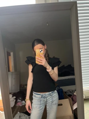 Svart volangtopp med markerade axlar - Säljer en svart topp med volangdetaljer vid axlarna. Toppen är ärmlös och har en tight passform som framhäver figuren. Perfekt för dig som vill ha en trendig och stilren look. Materialet känns mjukt och stretchigt.