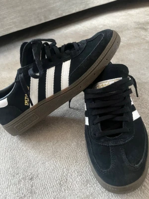 Adidas Handball Spezial svart/vit - Säljer ett par Adidas Handball Spezial sneakers i svart mocka med vita detaljer och gummisula. Klassisk Adidas-logga i guld på plösen och retroinspirerad design. Skorna har svarta snören och en låg silhuett som passar till många stilar.