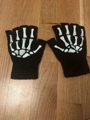Skelettmönstrade fingerlösa handskar - Svarta fingerlösa handskar med vitt skelettmönster på ovansidan. Perfekta för maskerad, cosplay eller halloween. Tillverkade i mjukt tygmaterial och ger en cool och edgy look till din outfit.