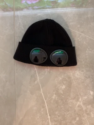 Svart mössa med glasögon C.P. Company - Säljer en svart ribbstickad mössa från C.P. Company med ikoniska inbyggda glasögon på uppviket. Mössan är gjord i mjukt material och har en klassisk form med bred kant. Perfekt för dig som vill sticka ut med en unik accessoar.