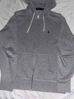 Grå hoodie från Polo Ralph Lauren - Grå zip-hoodie från Polo Ralph Lauren med vit dragsko i huvan och klassisk broderad logga på bröstet. Mjuk bomullskänsla, kängurufickor och ribbade muddar. Perfekt för en chill och stilren look.