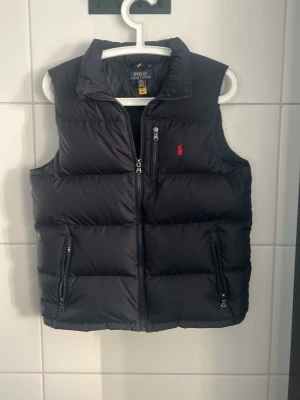 Rhalp laurent väst - Snygg svart dunväst från Polo Ralph Lauren med röd broderad logga på bröstet. Västen har hög krage, två dragkedjeförsedda fickor och quiltad design. Perfekt för lager på lager och håller dig varm med stil.