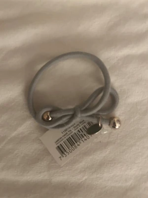 Grå hårsnodd med metallpärlor - Säljer en stilren grå hårsnodd från Jabon. Snodden har två silverfärgade metallpärlor och en liten metallbricka som detalj. Perfekt för att sätta upp håret på ett snyggt och enkelt sätt. Materialet är elastiskt tyg med metallinslag.