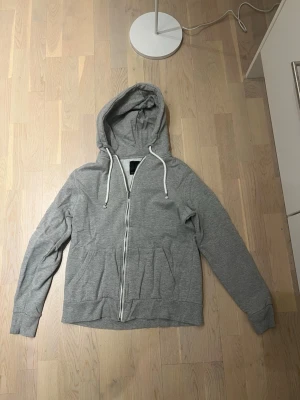 Grå zip hoodie med dragsko - Snygg gråmelerad hoodie från lager 157 med dragkedja framtill och vita dragsnören i huvan. Klassisk passform med fickor på sidorna och mjukt material som känns skönt mot huden. Perfekt för en avslappnad stil.