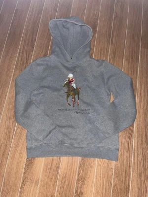 Grå hoodie Polo Bear Ralph Lauren - Snygg grå hoodie från Ralph Lauren med Polo Bear-tryck på bröstet där björnen rider häst och spelar polo. Klassisk huva och känguruficka framtill. Mjuk bomullskänsla och avslappnad passform, perfekt för en cool och sportig stil. Storlek XL (18-20) vilket motsvarar S. Bara hör av er vid funderingar!😃