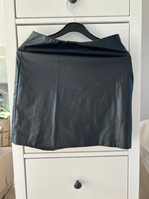 Svart kort skinnkjol - Snygg svart kort kjol i skinnimitation med enkel och stilren design. Kjolen har rak passform och är perfekt för att skapa en edgy look. Inga synliga detaljer eller fickor, vilket ger ett minimalistiskt intryck.