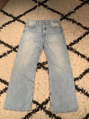 Acne Studios Jeans Light Blue W29 L32 Dam - Ett par ljusblå jeans från Acne Studios med utsvängda ben. Jeansen har en klassisk femficksmodell och knappgylf. De har en ljus tvätt med lätt slitna detaljer för en avslappnad look.