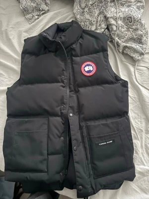 Svart dunväst från Canada Goose - Snygg svart dunväst från Canada Goose med klassisk logga på bröstet och två stora fickor framtill. Västen har hög krage, dragkedja och tryckknappar. Perfekt för lager-på-lager och håller dig varm med sin fluffiga dunfyllning.