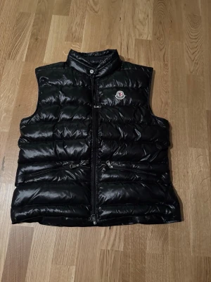 Svart dunväst från Moncler - Snygg svart dunväst från Moncler med glansig finish och klassisk logotyp på bröstet. Västen har hög krage, två fickor med dragkedja och quiltad design. Perfekt för lager-på-lager och håller dig varm under kyliga dagar.
