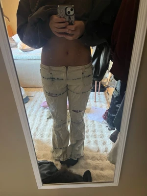 Beige slitna jeans med tie-dye detaljer - Säljer ett par beige/vita jeans med låg midja, vida ben och flera slitningar samt tie-dye detaljer i blått. Jeansen har dragkedja och knapp framtill och är perfekta för en trendig -look.