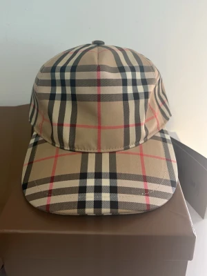 Burberry Keps - Säljer en klassisk keps från Burberry med ikoniskt rutmönster i beige, svart, vitt och rött. Kepsen har böjd skärm och justerbar passform. Perfekt för dig som vill ha en stilren accessoar med lyxig känsla och tidlös design.