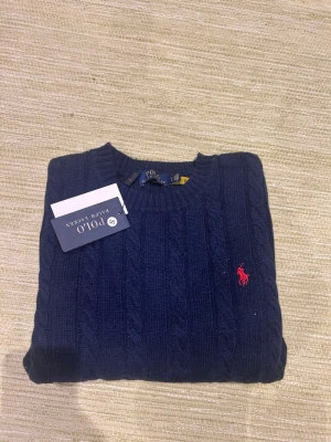Mörkblå kabelstickad tröja Polo Ralph Lauren - Kabelstickad tröja i mörkblått från Polo Ralph Lauren med klassisk röd logga broderad på bröstet. Rund halsringning och ribbade muddar vid ärmslut och nederkant. Perfekt för lager på lager och snygg till jeans. Tveka inte på att lägga bud!!