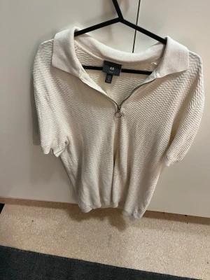 Vit kortärmad zip-tröja från H&M - Säljer en vit kortärmad t-shirt från H&M med slim fit passform. Tröjan har ett vågigt mönster i tyget, krage och en snygg dragkedja framtill med ringdetalj. Perfekt för dig som gillar stilrena plagg med en twist.