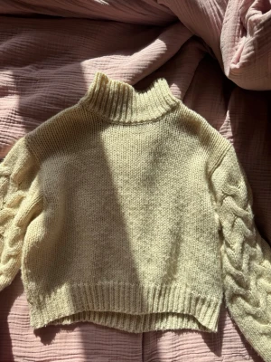 beige stickad tröja med flätmönster - Mysig beige stickad tröja med hög krage och breda ärmar med snyggt flätmönster. Tröjan har ribbstickade muddar vid ärmslut och nederkant samt en något boxig passform. Perfekt för dig som gillar en trendig och bekväm look.