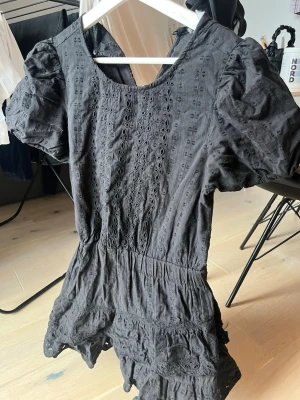 Svart broderad klänning från Zara - Supersnygg svart klänning från Zara med broderade detaljer och puffärmar. Kort modell med resår i midjan och volangkant nertill. Tillverkad i ett lätt tyg som känns luftigt och bekvämt. Perfekt för dig som vill ha en trendig och unik look.