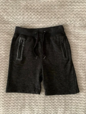 Svarta shorts med dragkedjor och snörning - Svarta shorts med elastisk midja, snörning och två fickor med dragkedjor framtill. Perfekta för chill dagar eller träning. Mjukt material som känns skönt mot huden och ger en avslappnad vibe.