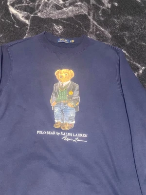 Mörkblå sweatshirt Polo Bear Ralph Lauren - Mörkblå sweatshirt från Ralph Lauren med ikoniska Polo Bear-trycket på bröstet. Rund halsringning och ribbade muddar vid ärmslut och nederkant. Perfekt för dig som gillar klassisk streetwear med en twist.