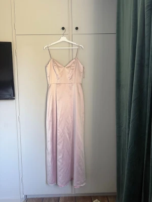 Ljusrosa långklänning i satin - Elegant ljusrosa långklänning med smala axelband och v-ringad urringning. Klänningen har en slät, glansig satinfinish och en figurnära passform som faller snyggt längs kroppen. Perfekt för bal eller andra festliga tillfällen. Är figursydd så den passar 38 eller 40 beroende på, enbart andvänd 1 gång.