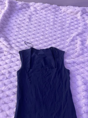 Marinblå ärmlös klänning - En stilren marinblå klänning med fyrkantig urringning och ärmlös design. Klänningen har en enkel, rak passform och är tillverkad i ett mjukt tyg som ger en klassisk look. Perfekt för dig som gillar minimalistisk stil.