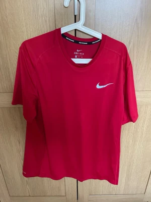 Röd Nike Dri-Fit löpartröja XL - Snygg röd löpartröja från Nike med Dri-Fit-teknologi som håller dig torr under träningen. Tröjan har rund halsringning, korta ärmar och en vit Nike-logga på bröstet. Perfekt för löpning eller gymmet.
