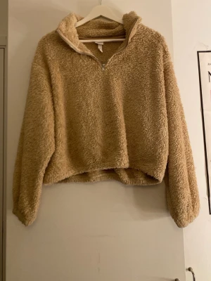 Beige teddyjacka med halv dragkedja - Mysig beige teddyjacka med halv dragkedja framtill och hög krage. Jackan har en avslappnad passform och är gjord i fluffigt, mjukt material som ger en riktigt skön känsla. Perfekt för kalla dagar när du vill vara både varm och snygg.
