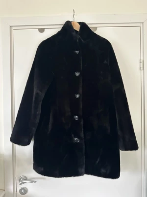 Svart fuskpäls kappa med knappar - RINO & PELLE- storlek S/M - Underbart mjukt svart jacka / kappa i högkvalitativt veganskt fuskpäls från RINO & PELLE. Stilren modell med knappar framtill och hög krage. Jackan har en rak passform och långa ärmar. Perfekt för att hålla värmen under kallare dagar. Det står storlek 36 (S) och är i oversize modell så passar även storlek 38 (M) i mer figurnära rak modell. Mycket gott skick, som ny!