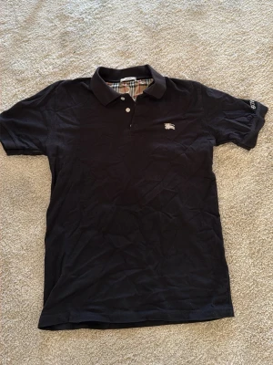 Svart pikétröja från Burberry - Svart pikétröja från Burberry med klassisk krage och tre knappar framtill. Ikoniskt rutigt mönster på insidan av kragen och diskret Burberry-logga på bröstet. Perfekt för en stilren och tidlös look. Pris kan diskuteras