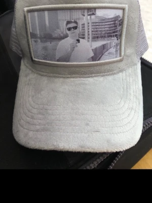 Grå meshkeps med fototryck - Cool grå keps med mesh på sidorna och baksidan. Framsidan har ett unikt fototryck i svartvitt som ger kepsen en personlig touch. Materialet är en blandning av mjuk mocka-liknande tyg och mesh, perfekt för dig som vill sticka ut.