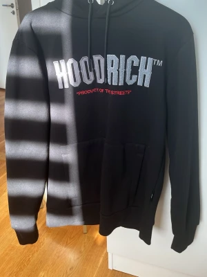 Svart HOODRICH hoodie med tryck - Svart hoodie från HOODRICH med stor vit logga och texten 'PRODUCT OF THE STREETS' i rött på bröstet. Klassisk känguruficka framtill, huva med dragsko och dragkedja på ärmen för extra stil. Perfekt för dig som gillar streetwear och vill sticka ut. 100% ÄKTA 