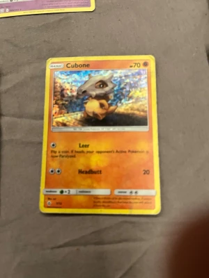 Pokémonkort Cubone holografisk - Säljer ett holografiskt Pokémonkort med Cubone-motiv. Kortet har en färgstark bakgrund i orange och gult, med Cubone i fokus. Text och symboler i svart och vitt, samt gul ram runt kortet. Perfekt för samlare eller Pokémonfans.