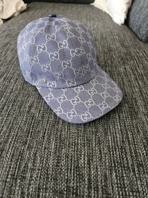 Blå keps från Gucci med GG-mönster - Snygg blå keps från Gucci med klassiskt GG-monogram över hela kepsen. Kepsen har böjd skärm och är tillverkad i ett tåligt tygmaterial. Perfekt för dig som vill sticka ut med en lyxig accessoar.