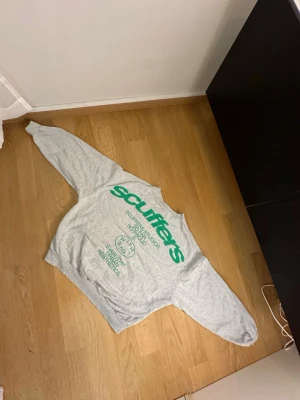 Grå sweatshirt från Scuffers - Grå oversized sweatshirt från Scuffers med stor grön logga och tryck framtill. Tröjan har rund halsringning, långa ärmar och ribbade muddar vid ärmslut och nederkant. Perfekt för en avslappnad streetwear-look. Tröjan är i storlek S men passar även M