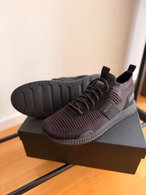 Svarta sneakers från BOSS - Stilrena svarta sneakers från BOSS med stickad ovandel och diskreta logodetaljer på sidan och plösen. Skorna har en chunky sula och elastisk passform med snörning. Perfekta för en modern och sportig look.