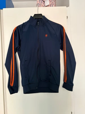 Blå träningsjacka från Skill - Mörkblå träningsjacka från Skill med dragkedja och två fickor framtill. Snygga orange ränder längs ärmarna och liten logga på bröstet. Perfekt för träning eller sportiga dagar. Tillverkad i ett mjukt och stretchigt material.