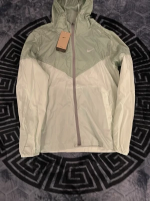 Nike Miller  - Pris kan diskuteras. Snygg ljusgrön vindjacka från Nike med dragkedja framtill och huva. Jackan har tvådelad färgton i mintgrönt och ljusgrönt, samt reflekterande Nike-logga på bröstet. Tillverkad i lätt och tunt material, perfekt för blåsiga dagar. Praktiska sidofickor och justerbar huva.