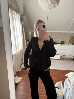 Svart satinblus med v-ringning - Snygg svart blus i glansigt satinmaterial med djup v-ringning och långa ärmar. Blusen har en avslappnad passform och stilrena detaljer vid ärmsluten. Perfekt att styla med både jeans och kjol för en trendig look.