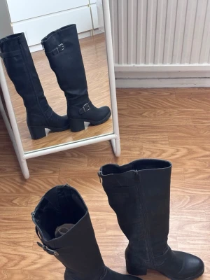 Svarta höga boots med spännen - Säljer ett par svarta höga boots i syntet med chunky klack. De har en del defekter, vissa platser har ”lädret” skavts av. Skorna är ganska gamla men har inte använts så mycket. Slutar precis under knäna. 