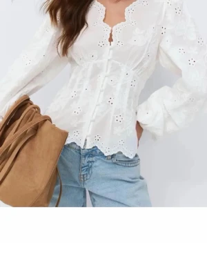 Vit broderad blus med knappar - Superfin vit blus med broderade detaljer och dekorativa knappar framtill. Blusen har långärmade puffärmar, v-ringning med vågig kant och är gjord i ett lätt bomullstyg. Perfekt att styla med jeans för en trendig look.