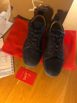 Christian Louboutin svarta mockasneakers - Säljer ett par svarta sneakers från Christian Louboutin i lyxig mocka med ikonisk röd sula och broderad logga på hälen. Skorna har snörning och rund tå, och kommer med originalkartong, dustbag och äkthetskort. Perfekta för dig som vill sticka ut med exklusiv stil.