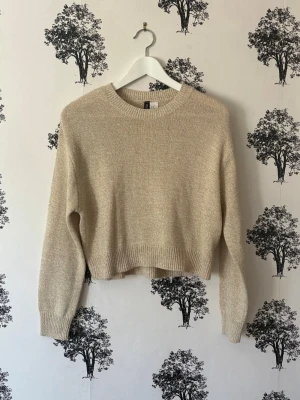 Beige stickad tröja från H&M - En enkel och snygg beige stickad tröja från H&M med rund halsringning och ribbade muddar. Tröjan har en något kortare passform och långa ärmar, perfekt för lager-på-lager. Materialet är mjukt och behagligt.