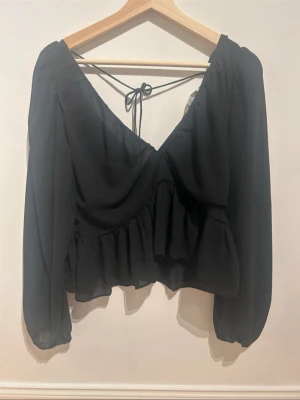 Svart blus med v-ringning och volang - Snygg svart blus med djup v-ringning och knytband i ryggen. Blusen har volangdetaljer och är tillverkad i ett tunt, flowy material som ger en luftig känsla. Långärmad modell med ballongärmar och avslappnad passform.