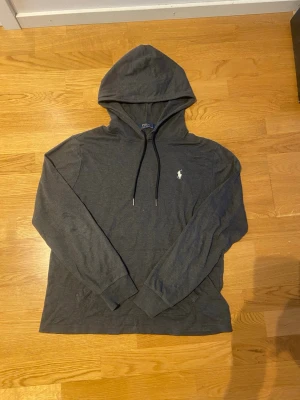 Mörkgrå hoodie från Polo Ralph Lauren - Snygg mörkgrå hoodie från Polo Ralph Lauren med klassisk huva och dragsnören. Ikonisk vit logga broderad på bröstet. Långärmad och tillverkad i mjukt material som är riktigt skönt. Perfekt vardags hoodie!