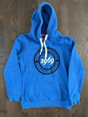 La Folie Douce Hoodie Herr - Cool blå hoodie med stor huva och vitt snöre. Tryck på bröstet med texten 'La Folie Douce 1969' och LFD-brodyr på ryggen. Mjuk bomullskänsla och klassisk känguruficka framtill. 