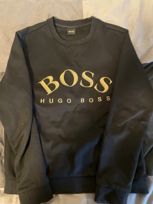 Svart sweatshirt från Hugo Boss - Cool svart sweatshirt från Hugo Boss med stort guldigt BOSS-tryck på bröstet. Tröjan har rund hals, ribbade muddar och är tillverkad i mjukt material som känns skönt mot huden. Perfekt för dig som gillar stilrena och trendiga plagg.