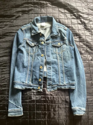 Klassisk blå jeansjacka - Snygg blå jeansjacka med guldfärgade knappar och två bröstfickor med lock. Jackan har en tidlös design med raka sömmar och krage. Perfekt att slänga över en hoodie eller t-shirt för en avslappnad look. Kvinnlig storlek 40